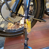 A close-up of a motorcycle's rear wheel and suspension with a black ratchet strap and gold-toned quick-release buckle secured across the wheel area, set on a wooden deck.